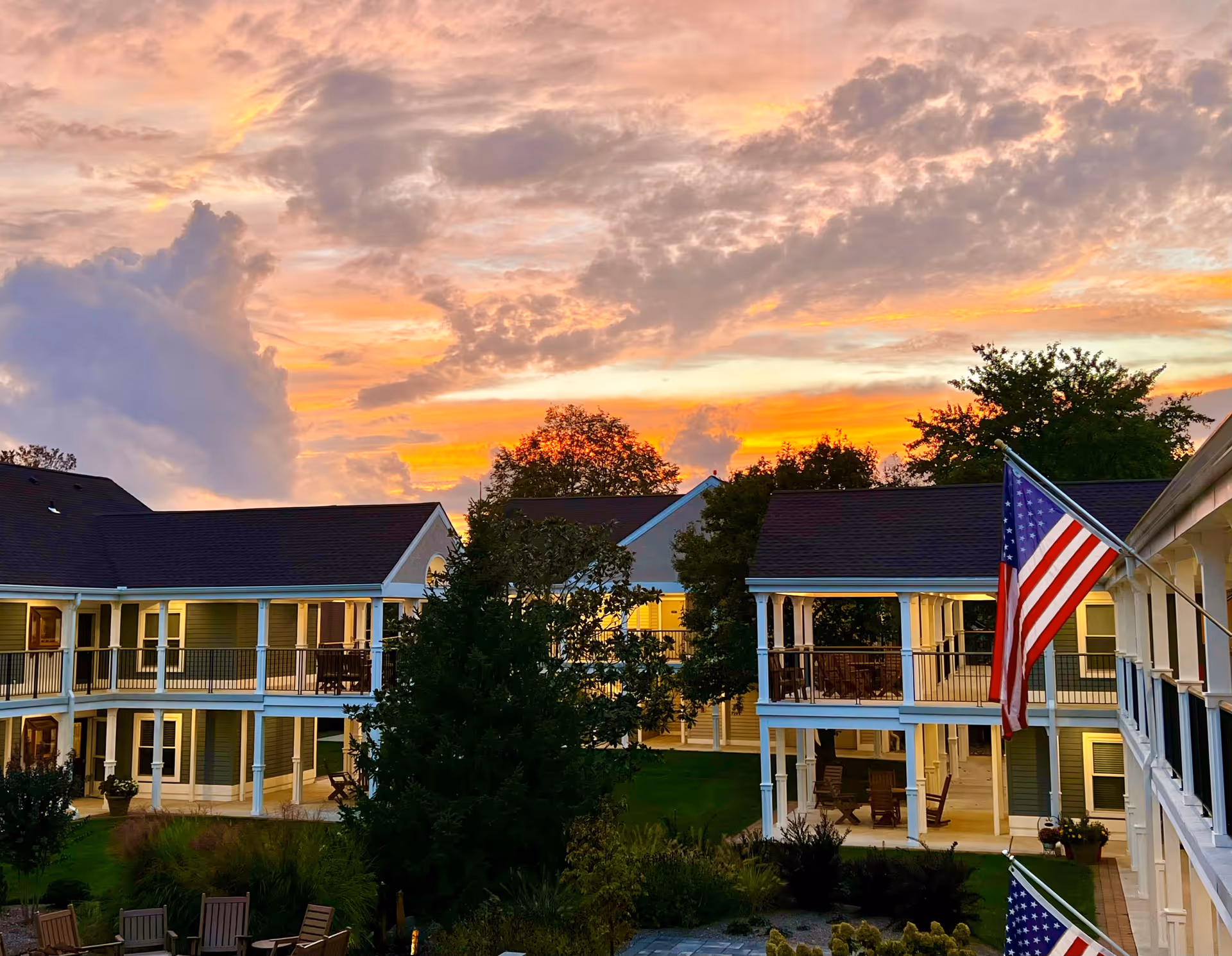 View of a senior living facility courtyard at sunset with two-story buildings featuring balconies and white railings, an American flag, trees, and outdoor seating areas.