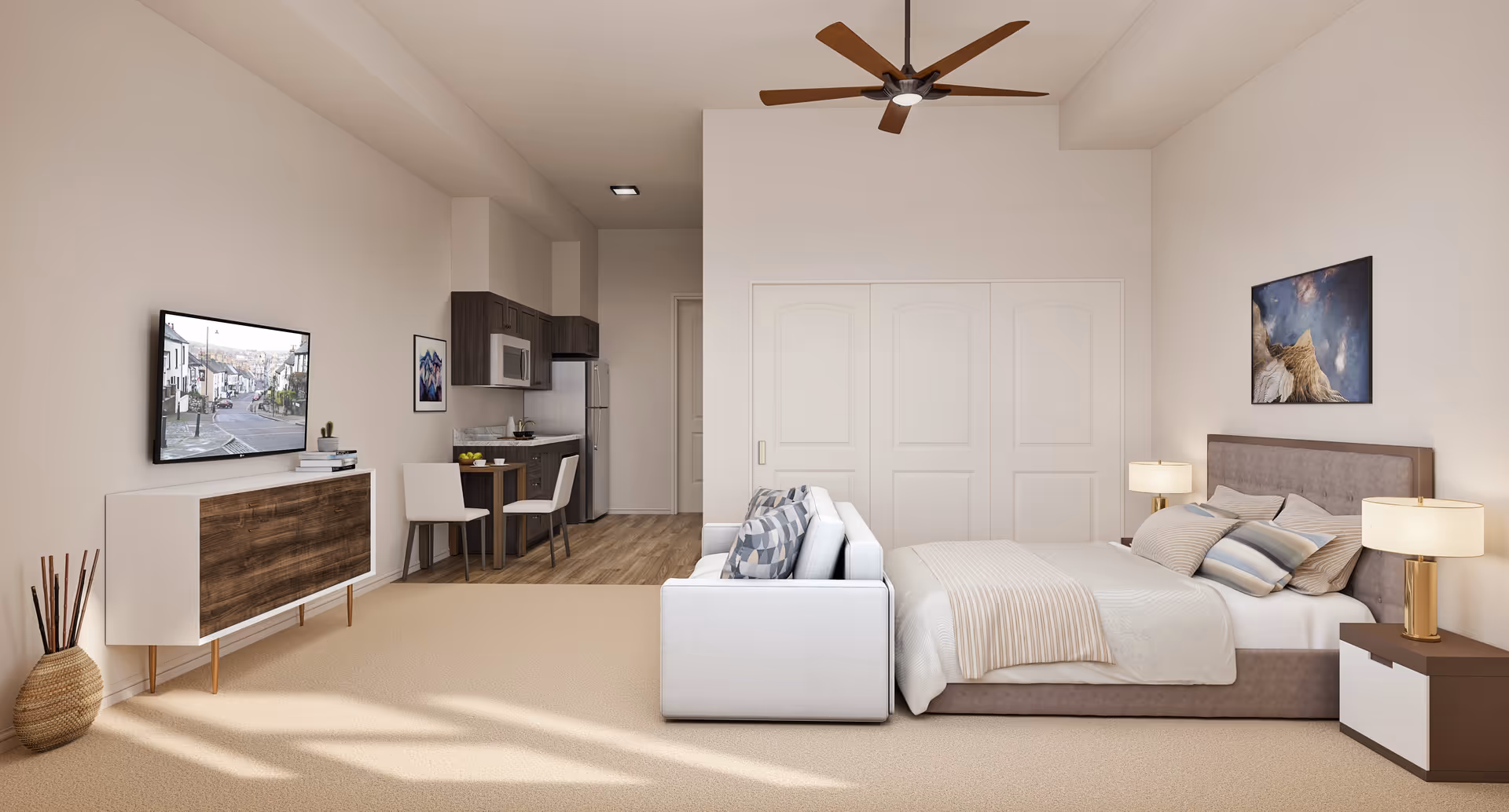 A modern studio apartment featuring a bed with multiple pillows and a beige blanket, two bedside tables with lamps, a white sofa facing a wall-mounted TV, a wooden cabinet, and a small dining table with two chairs. The kitchenette includes dark wood cabinets, a microwave, and a refrigerator. The room has beige carpeting, light-colored walls, a ceiling fan with wooden blades, and a framed picture above the bed.