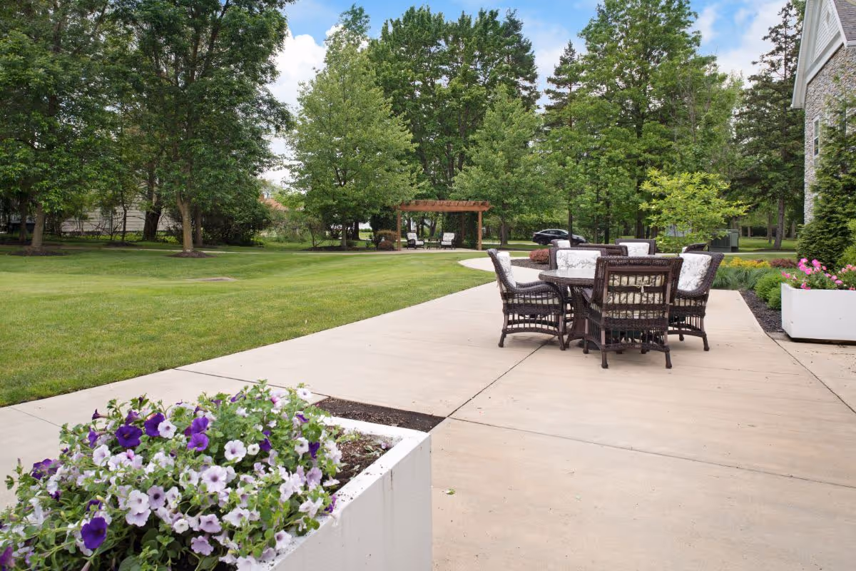 Outdoor patio area with a round table and six wicker chairs with cushions, surrounded by green grass, trees, and flower planters. A wooden pergola with two chairs is visible in the background under a partly cloudy sky.
