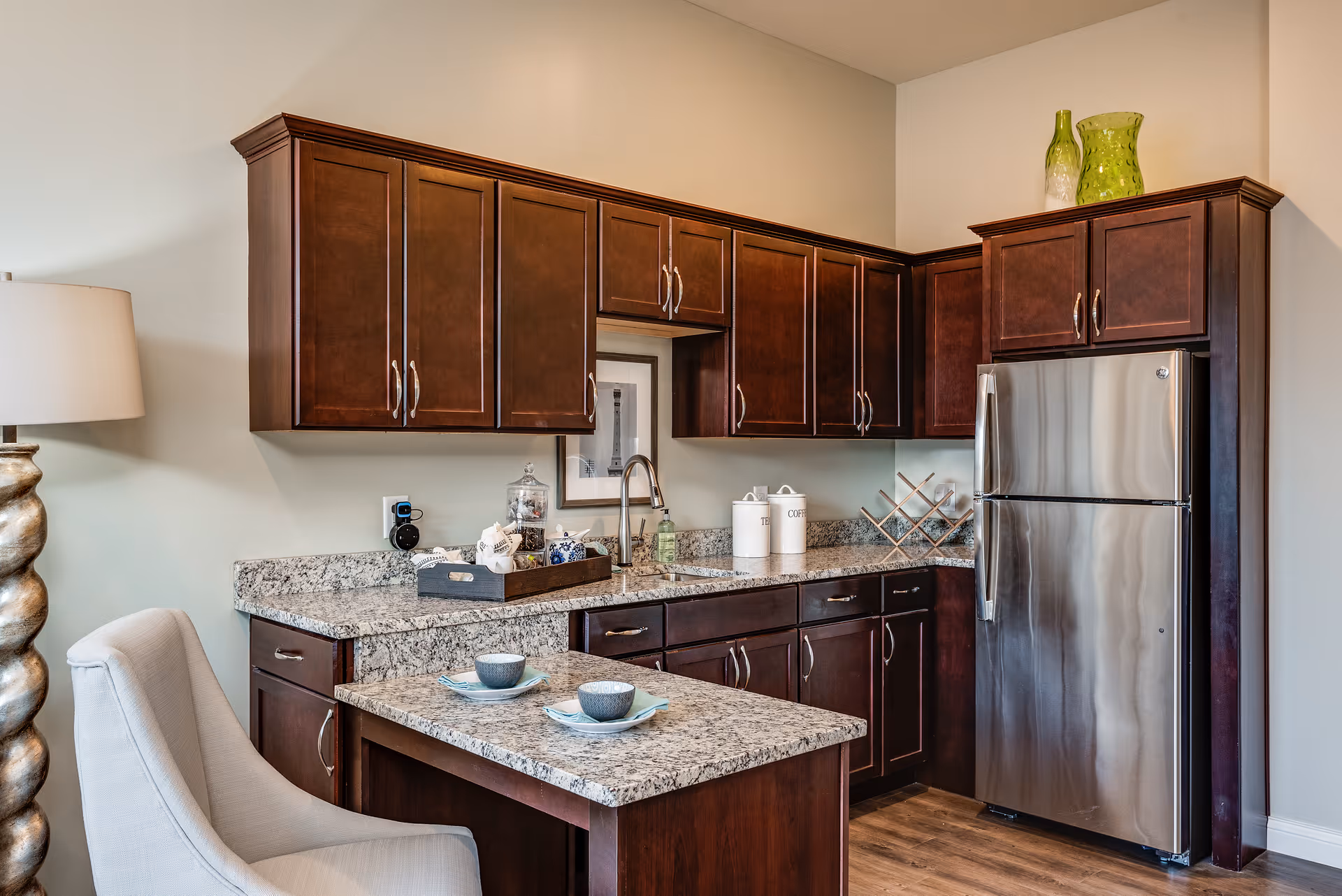 A modern kitchen with dark wooden cabinets, granite countertops, and a stainless steel refrigerator. There is a small island with two place settings including bowls and plates. A decorative lamp and a cushioned chair are visible on the left side. The kitchen also features a sink, a framed picture on the wall, and decorative jars labeled 'Tea' and 'Coffee'.