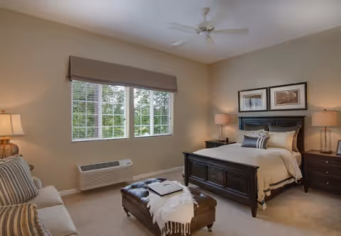 Bright, neatly decorated bedroom with a dark wood bed, matching nightstands and lamps, a sofa, ottoman and a window with blinds.