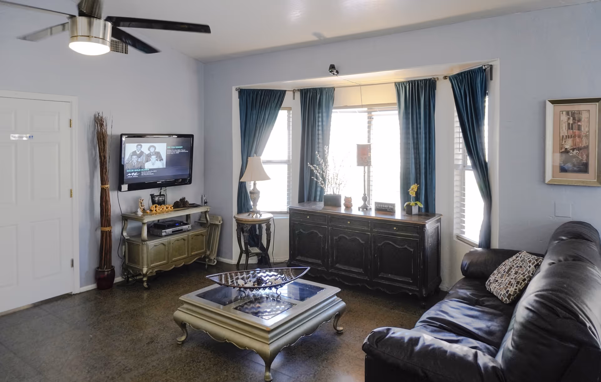 A cozy living room with a black leather sofa on the right, a decorative coffee table in the center, and a TV mounted on the wall to the left. The room has light blue walls, a ceiling fan with a light, and three windows with blue curtains letting in natural light. There is a wooden sideboard under the windows with decorative items and a lamp on a small round table next to it.