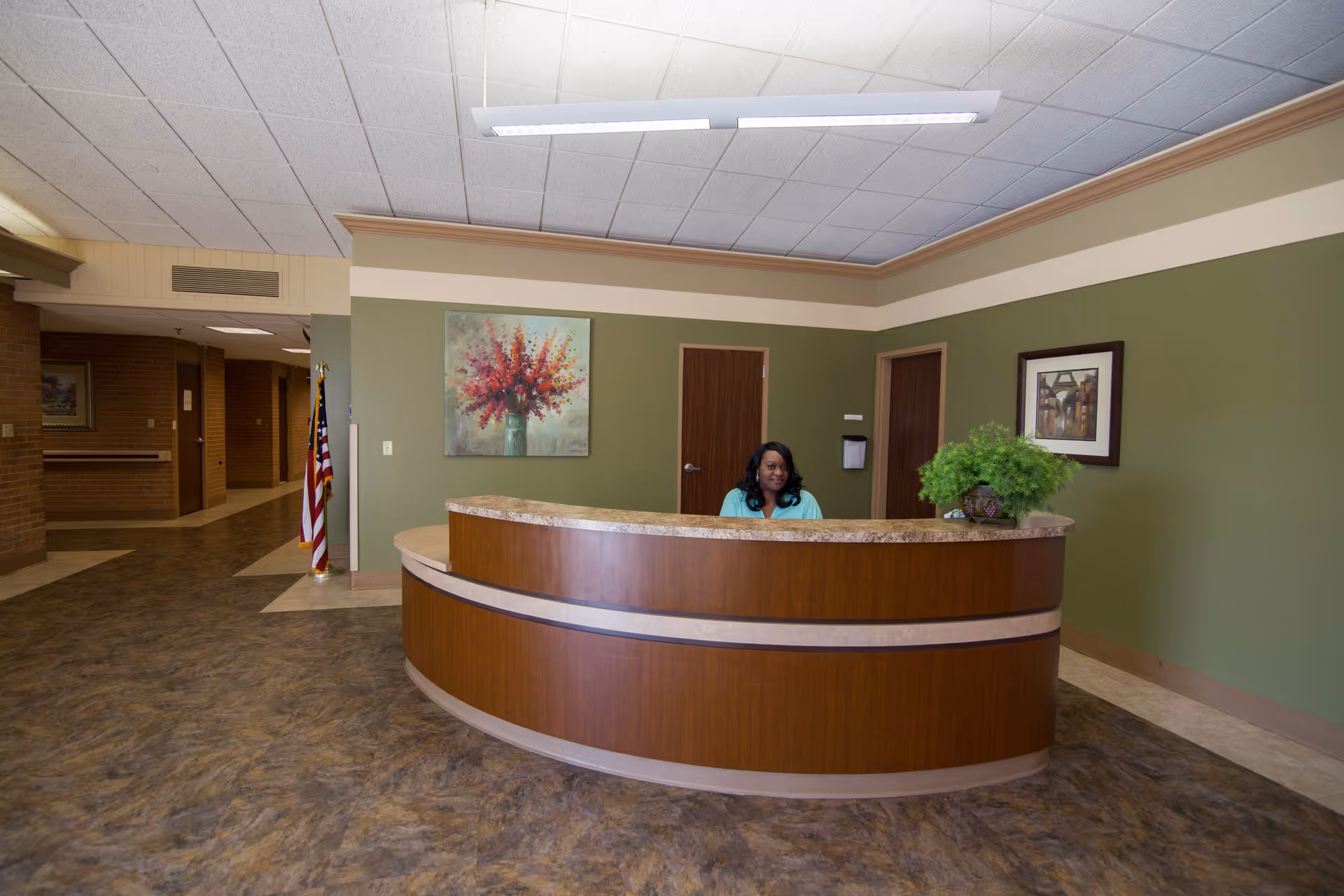 Reception area in a senior living facility with a curved wooden desk, a woman sitting behind the desk, green walls decorated with framed artwork, and an American flag in the background.