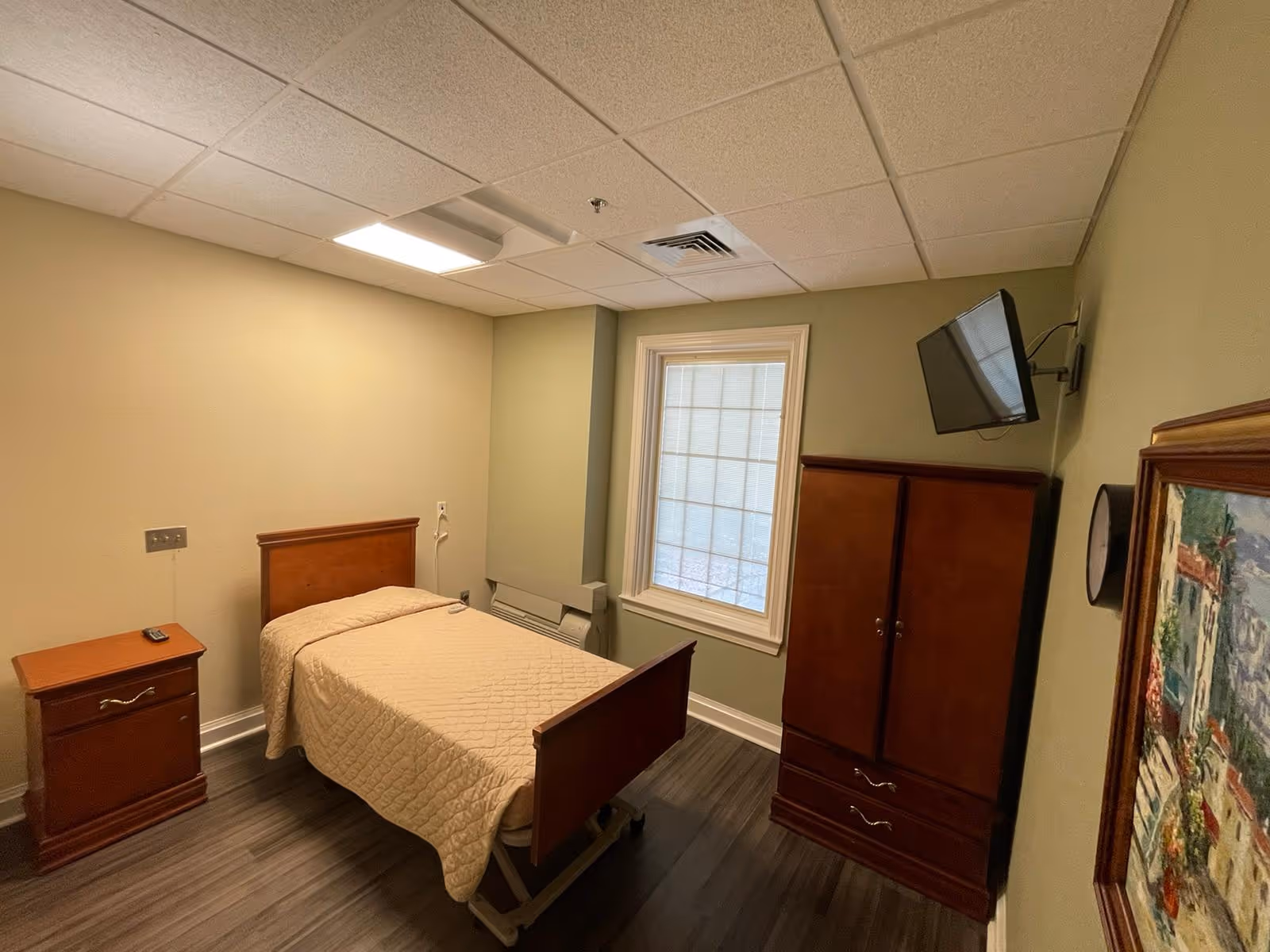 A small, simple bedroom in a senior living facility with a single bed covered in a beige quilt, a wooden nightstand with a remote control on it, a wooden wardrobe, a wall-mounted flat screen TV, a window with blinds, and a framed painting on the wall.
