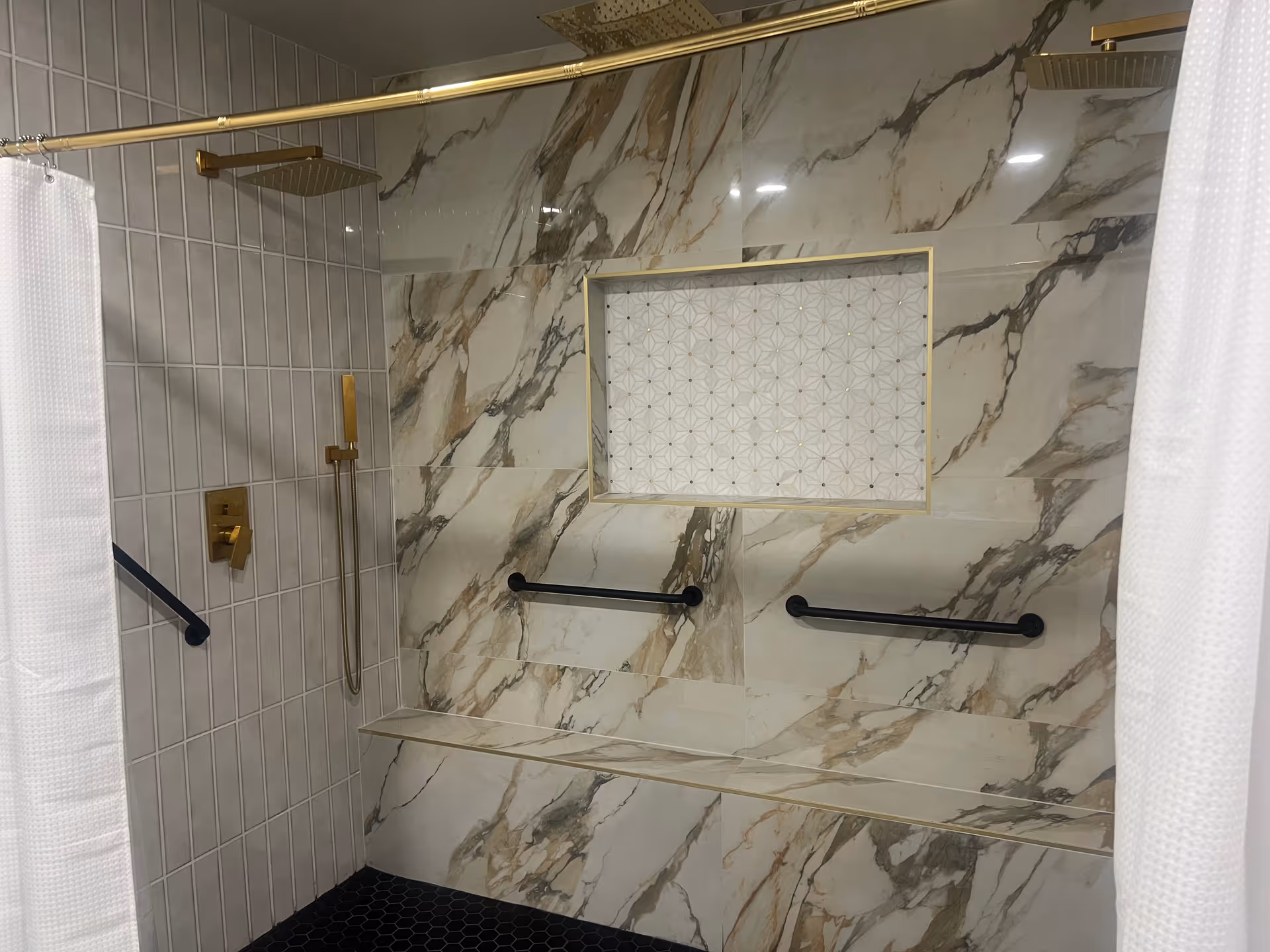 Marble-tiled accessible shower with built-in bench, black grab bars, gold shower fixtures, and white curtains.