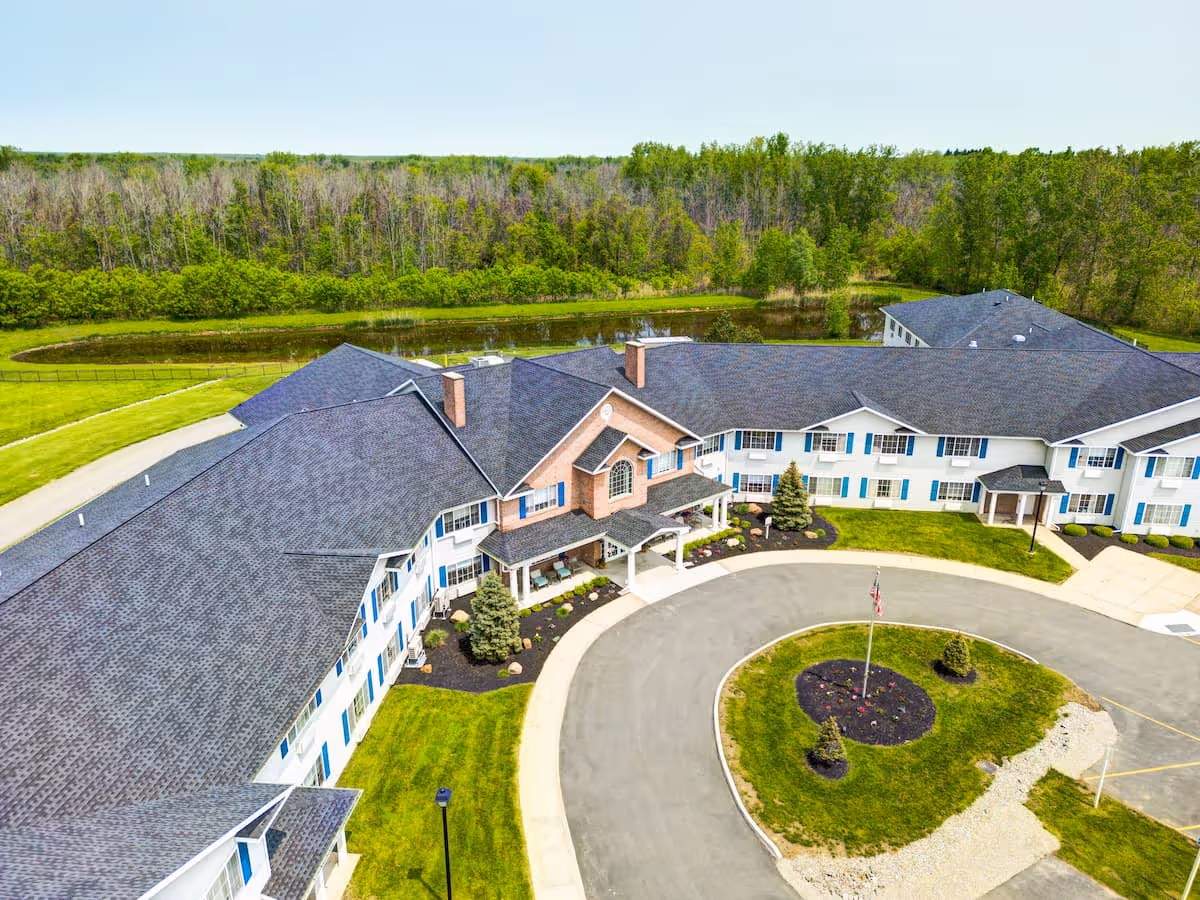Aerial view of Evergreen Place, a large senior living facility with a U-shaped building surrounded by well-maintained green lawns, a circular driveway with a flagpole in the center, and a wooded area in the background.
