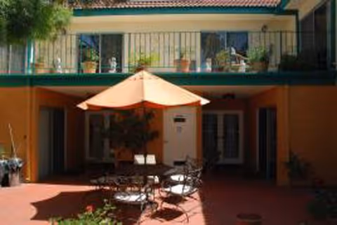 Outdoor courtyard area with a round table and chairs under a large beige umbrella, surrounded by a two-story building with balconies and potted plants.