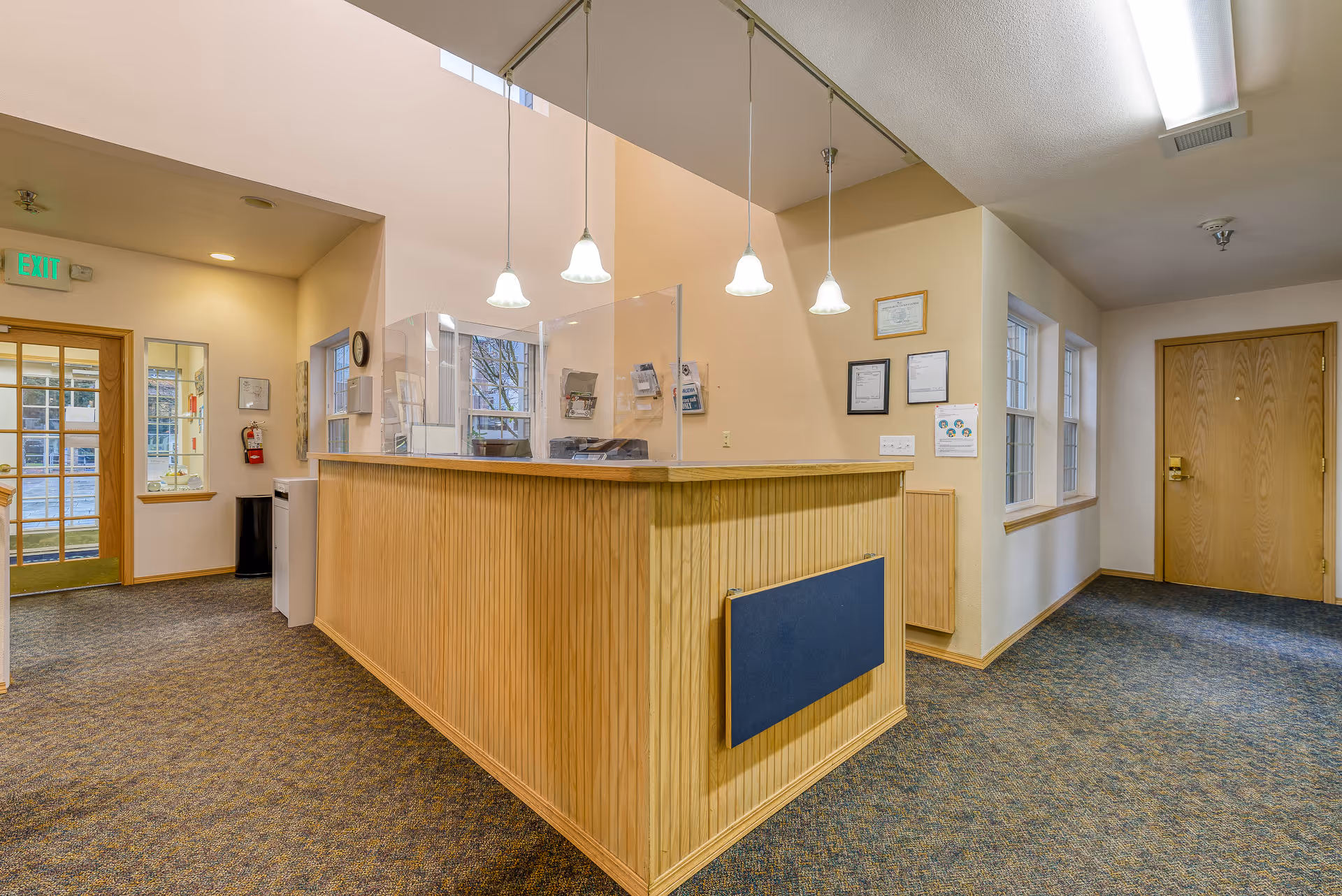 Reception area inside Hearthside Manor Memory Care with a wooden front desk featuring a clear protective screen. The area is well-lit with hanging pendant lights and has carpeted flooring. There are windows on the right wall and a door at the end of the hallway. Exit sign and fire extinguisher are visible near the entrance door on the left.