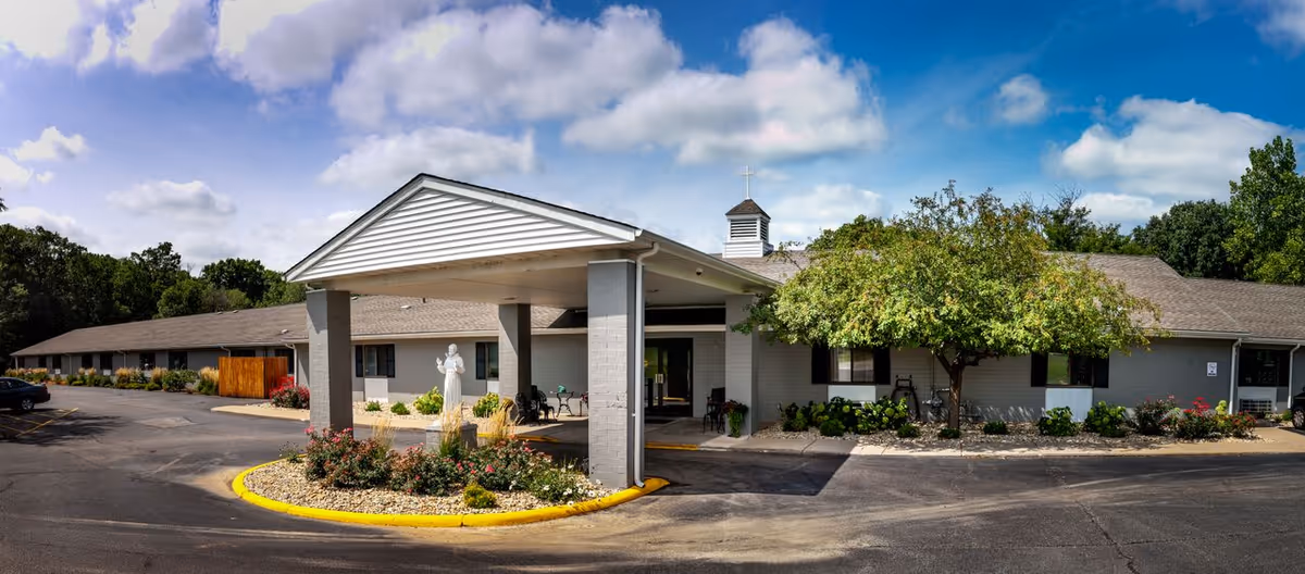 Front exterior of the St. Francis Woods senior living building with a covered entrance, circular flower bed and statue.