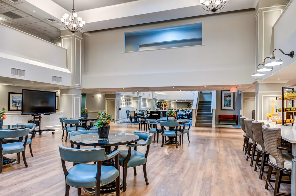 Spacious and well-lit common area with round tables and blue cushioned chairs, a large flat-screen TV on a stand, a bar counter with high chairs, and a staircase leading to an upper level. The room features wooden flooring, modern chandeliers, and decorative plants on tables.