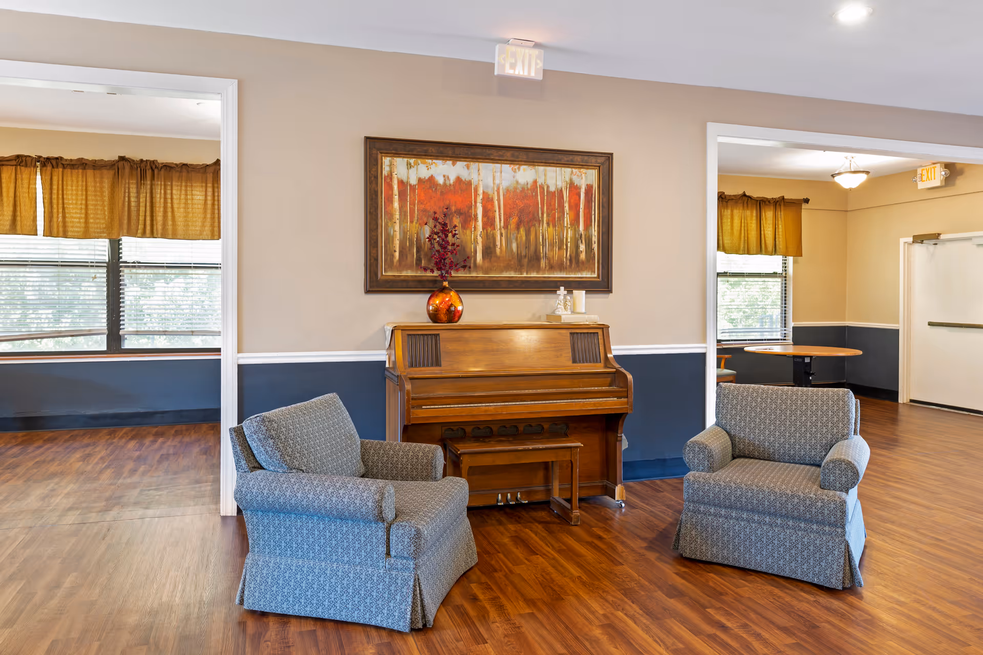Two upholstered chairs flank a wooden piano under framed artwork in a communal seating area.