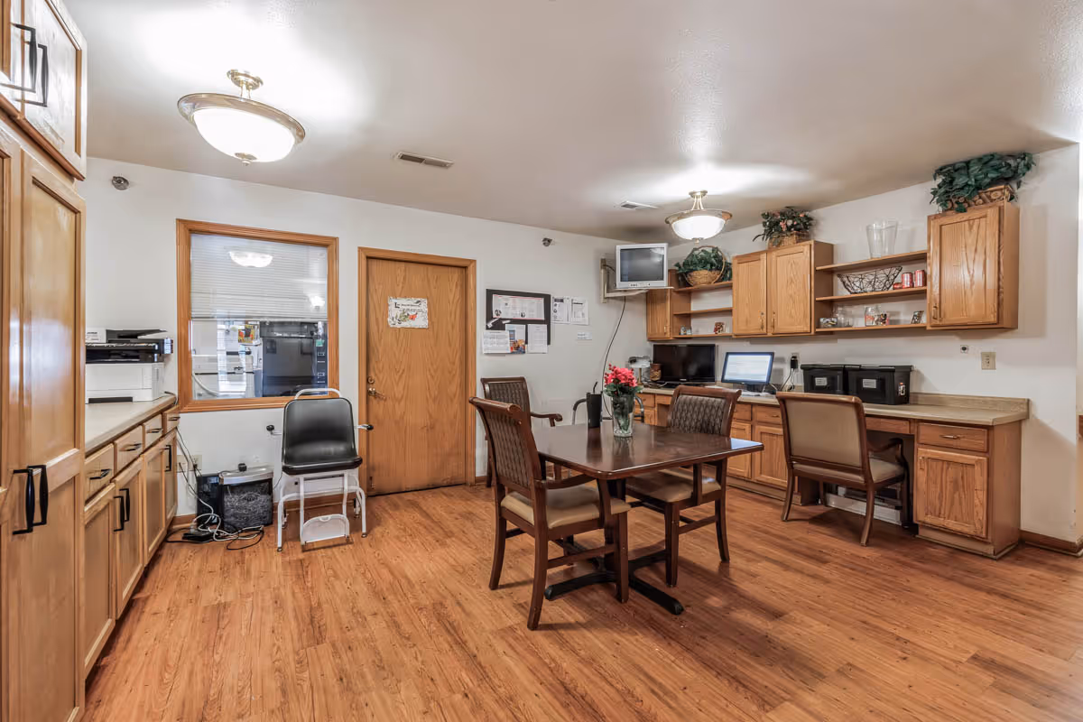 Interior room with wooden flooring and cabinetry, featuring a table with four chairs in the center, a computer workstation with two chairs along the wall, a small TV mounted in the corner, and various office supplies and decorations on shelves and counters.