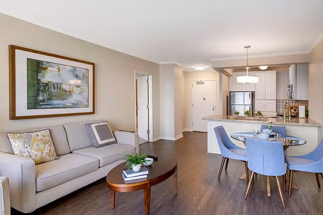 Open-plan living room and kitchen with a beige sofa, wooden coffee table, round dining table with blue chairs, and stainless-steel appliances.