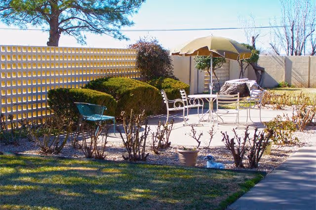 Outdoor patio area with metal chairs and a table under a large umbrella, surrounded by bushes and a decorative concrete block wall, with a grassy lawn and a small dog lying on the ground.