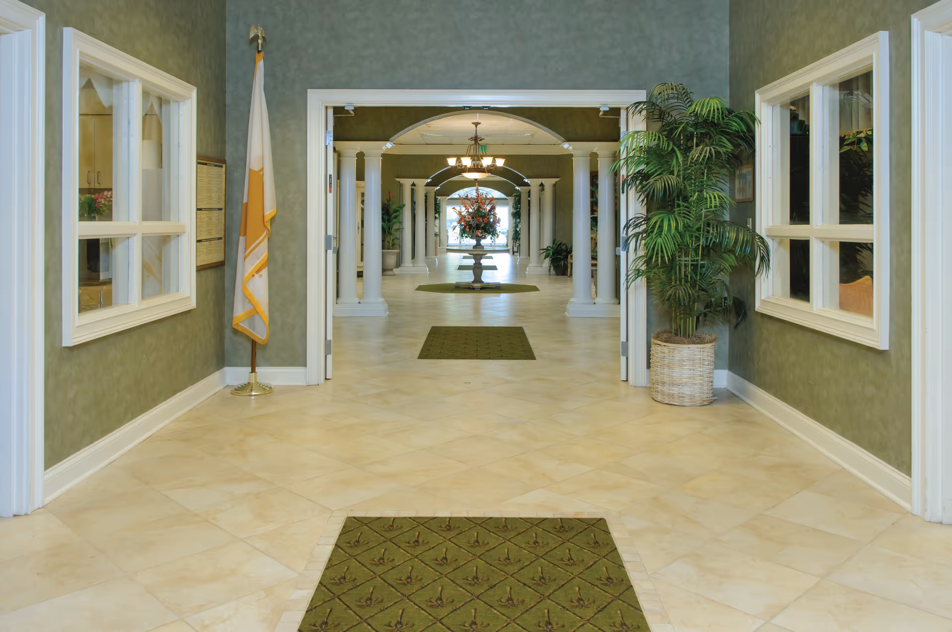 Well-lit lobby hallway with tiled floors, decorative columns, green rugs, potted plants, and a floral arrangement down the corridor.