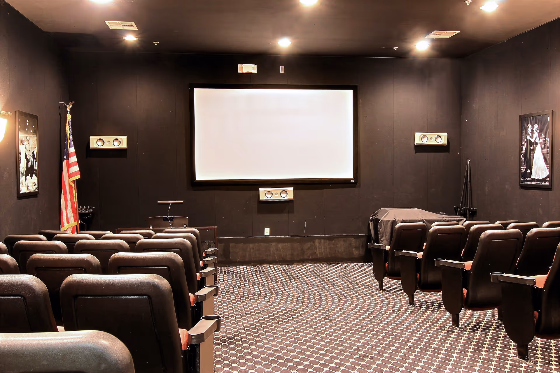 Interior view of a small theater room with rows of black cushioned seats facing a large blank projection screen. The walls are dark, and there are speakers mounted on the walls around the screen. An American flag stands in the corner, and framed black and white photos hang on the walls.