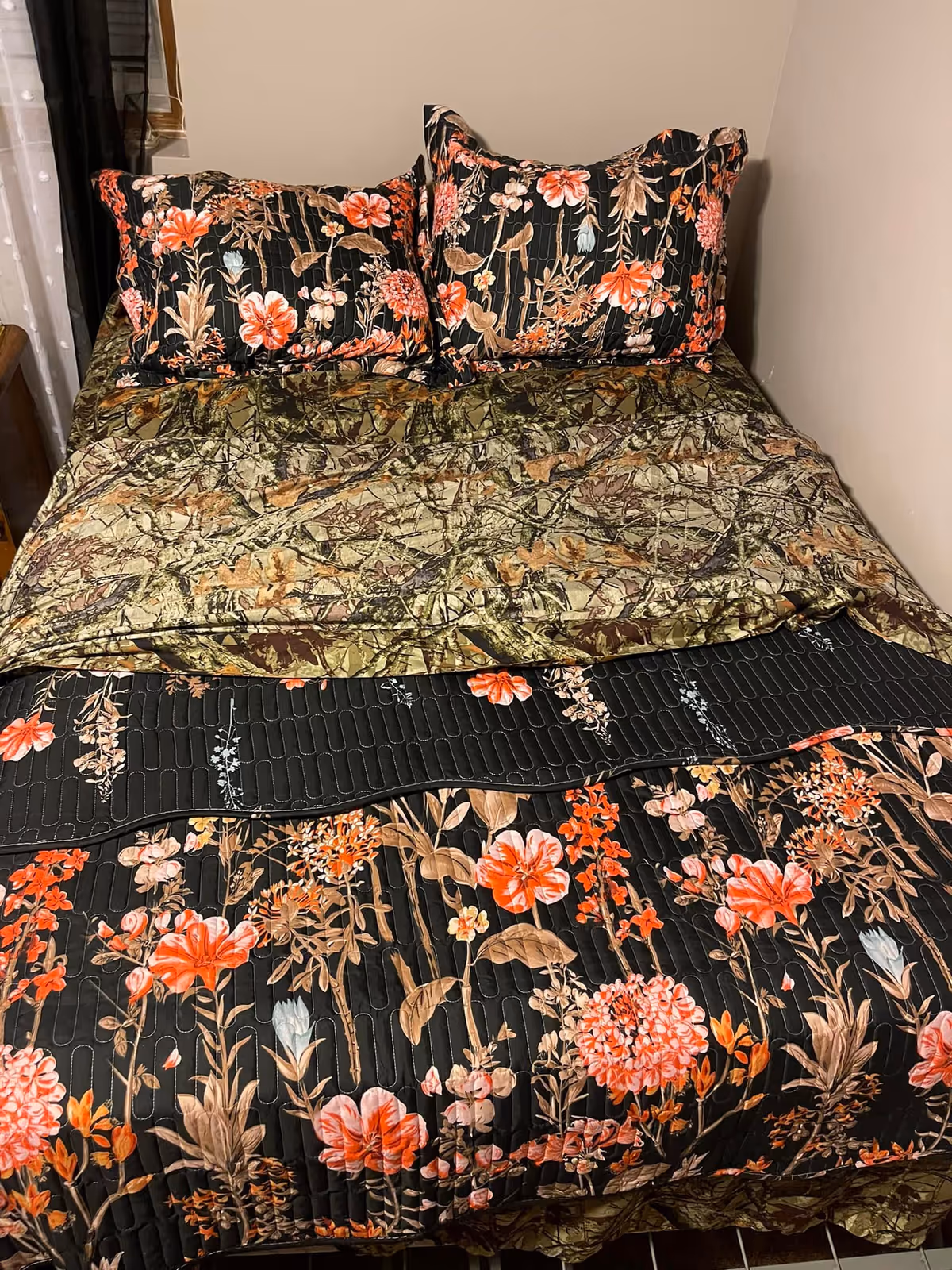 A neatly made bed with floral patterned pillows and a matching floral bedspread in shades of black, orange, and brown. The bed is placed against a beige wall with a window partially visible on the left side.
