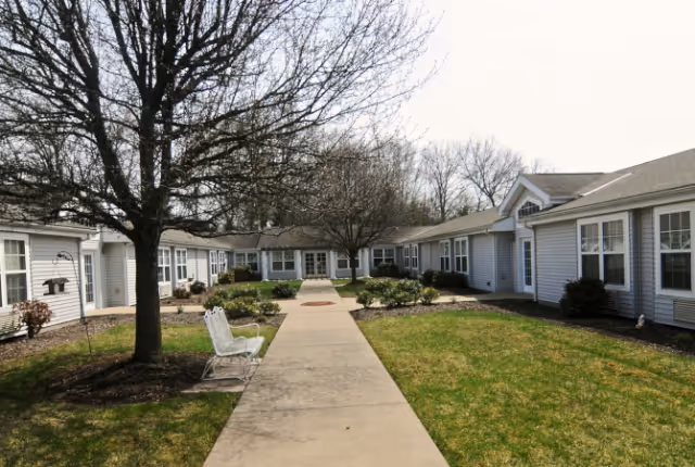 Outdoor courtyard area at The Addison of Lowrie Place featuring a concrete walkway lined with grass and small bushes, surrounded by single-story buildings with white siding and multiple windows. There are leafless trees and a white bench along the walkway.