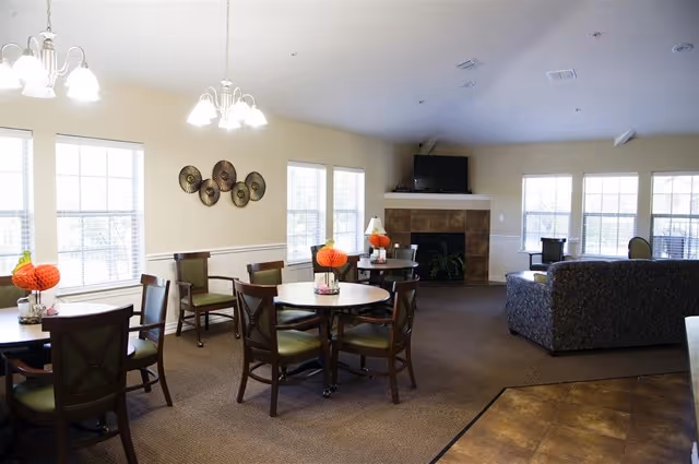 Bright communal dining and lounge area with round tables and chairs, a fireplace with a TV, and large windows.