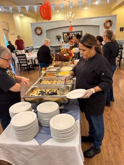 A woman in a black chef's coat serving food from a buffet table with trays of various dishes, including a blueberry-topped dessert. Several people are standing around the buffet and in the background, a dining area is visible with tables and chairs. The room is decorated with autumn wreaths and hanging banners.