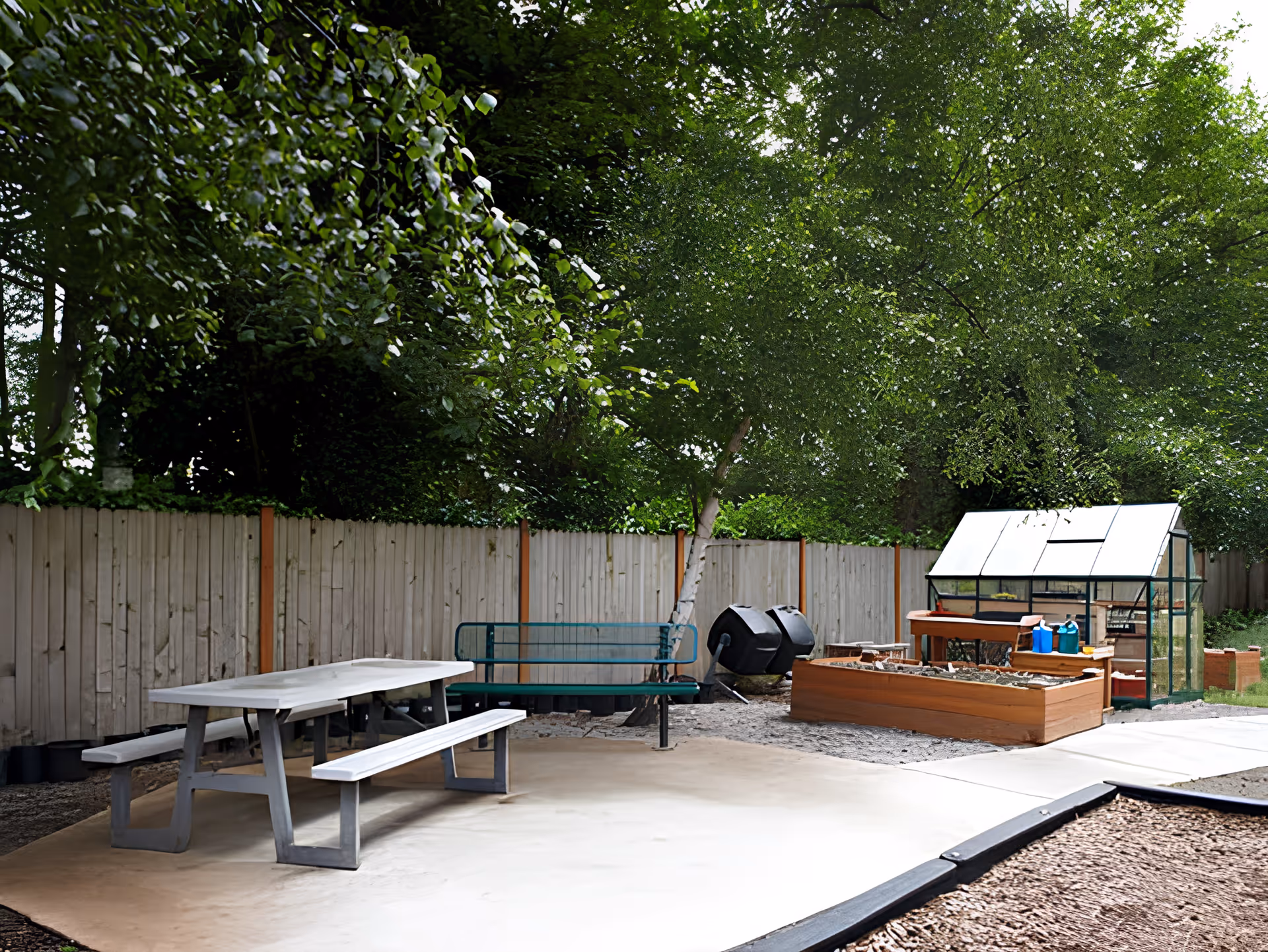 Outdoor garden area with a picnic table and benches on a concrete patio, a green metal bench, a tree, a wooden raised garden bed, a small greenhouse, and two black compost bins, all surrounded by a wooden fence and lush green trees.