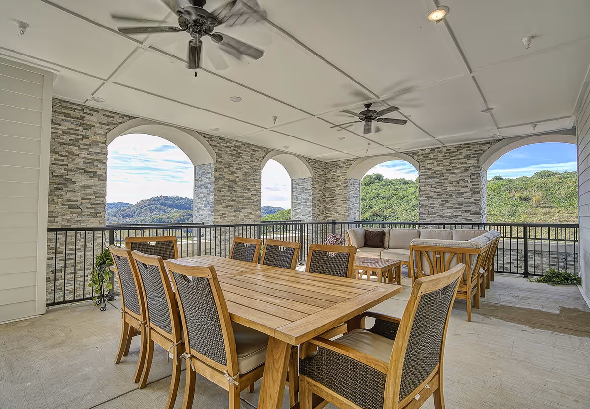 Covered outdoor patio with a wooden dining table, chairs and a cushioned seating area beneath arched openings overlooking green hills.