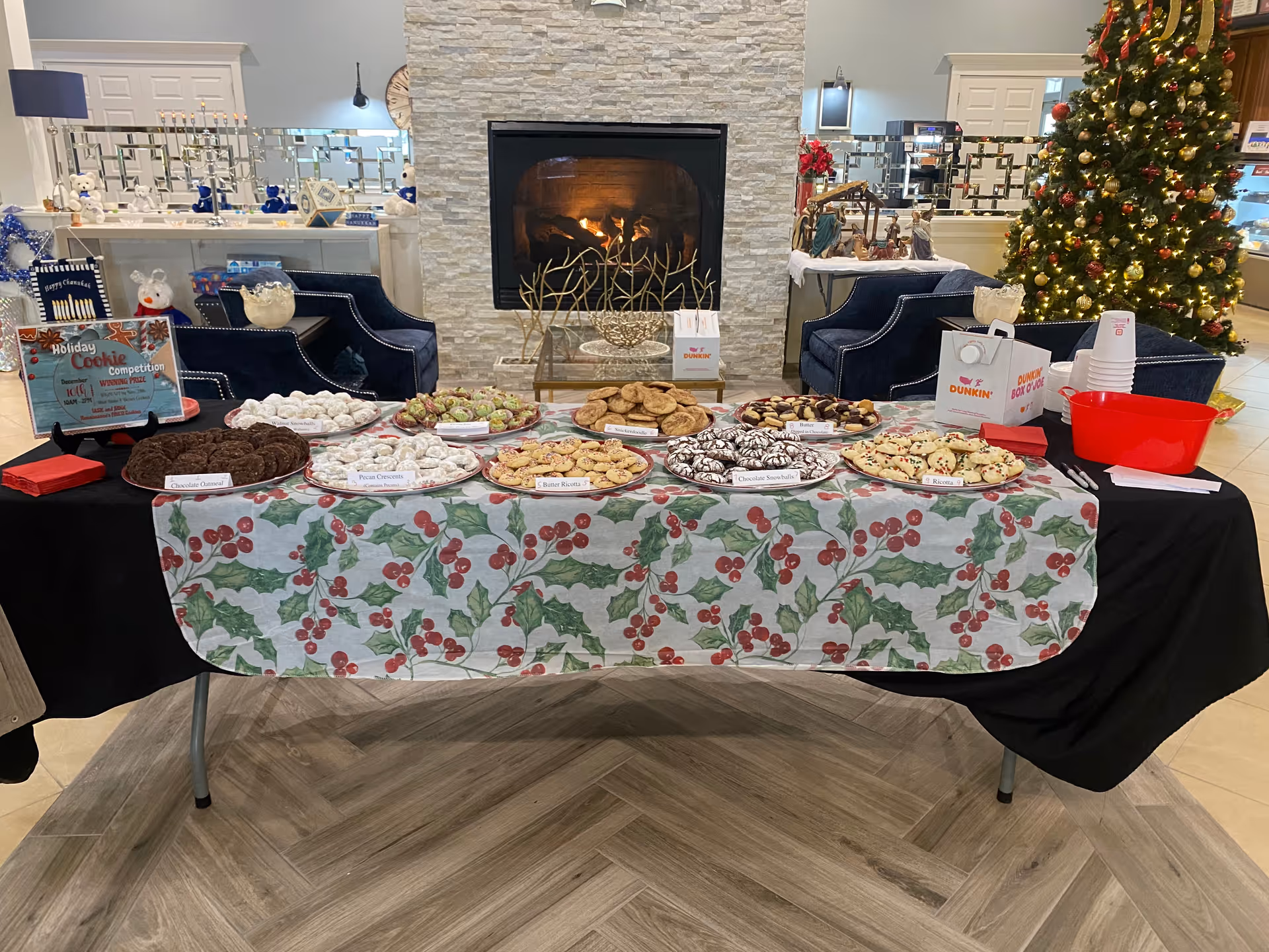 Festive table of assorted cookies displayed in front of a lit fireplace and a decorated Christmas tree in a facility lobby.