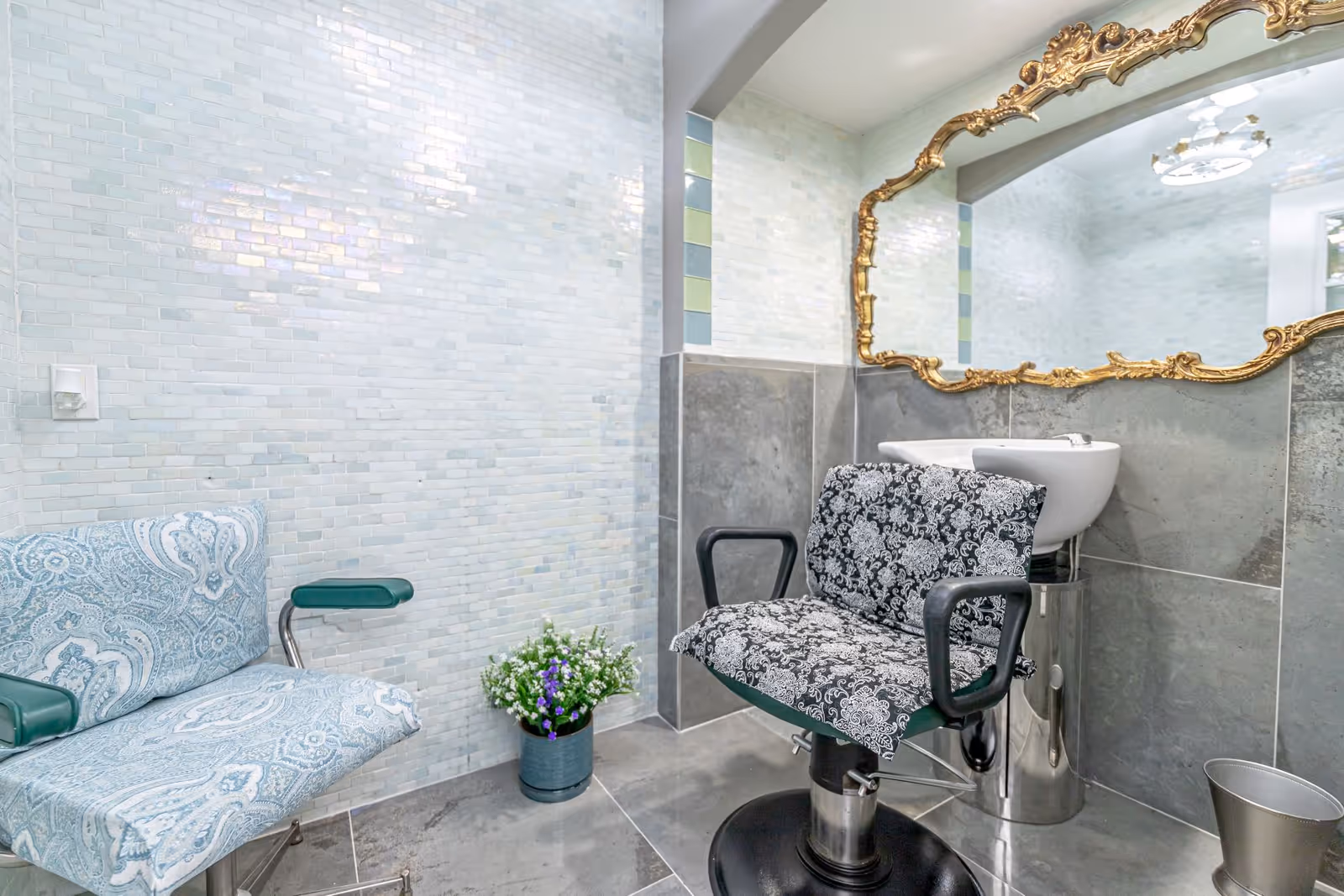 Interior of a salon area with a patterned salon chair in front of a wash basin, a blue patterned chair, a small potted plant with white and purple flowers, and a large ornate gold-framed mirror on the wall.