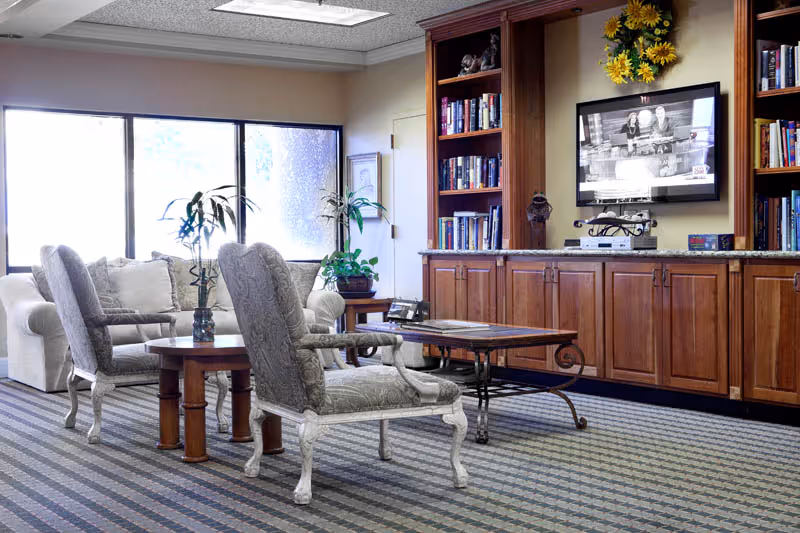 A cozy living room area with two upholstered armchairs and a sofa arranged around a wooden coffee table. The room features a large built-in wooden bookshelf and cabinet unit with a flat-screen TV mounted above it. There are decorative plants and a wreath with yellow flowers on the wall above the TV. The room has large windows letting in natural light and a patterned carpeted floor.