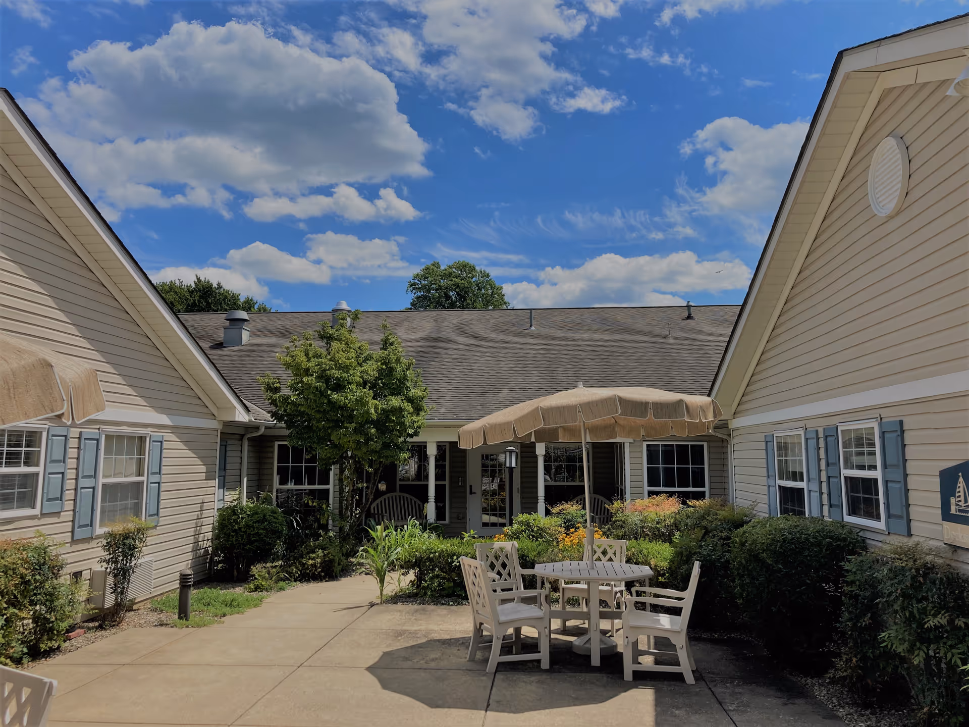 Outdoor courtyard area of a senior living facility with beige siding buildings on either side, a concrete walkway, green bushes, a small tree, and a round table with four chairs under a beige umbrella. The sky is blue with scattered clouds.
