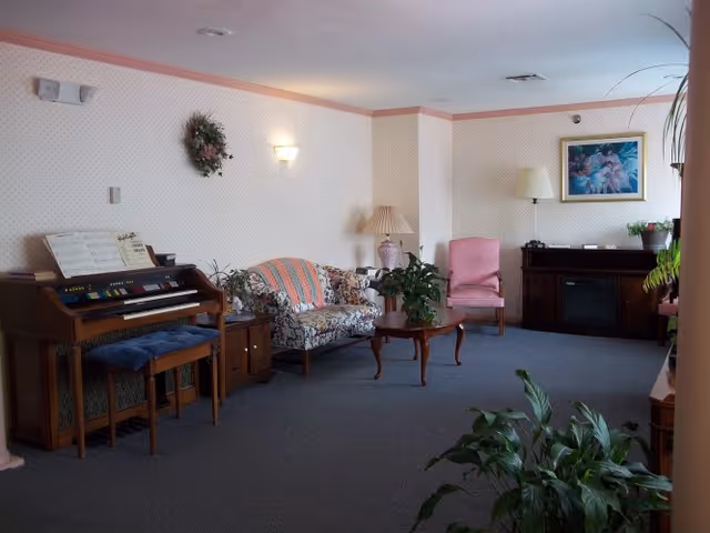 A cozy living room with a floral patterned loveseat, a pink armchair, a wooden coffee table, and an electric fireplace with a lamp and framed artwork above it. There is an organ with sheet music on the left side and several green plants around the room. The walls have light wallpaper with a subtle pattern and a pink border near the ceiling.