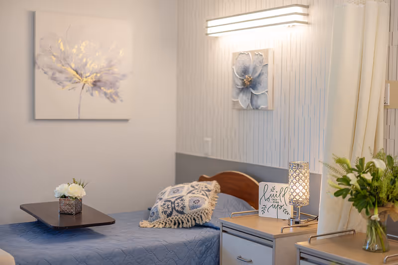 A neatly made single bed with a blue quilt and decorative pillow in a softly lit room. There is a small table with a flower arrangement on the bed, a bedside table with a lamp, a small sign that reads 'Be still each moment,' and a vase with green plants. The walls have floral artwork and a modern light fixture.