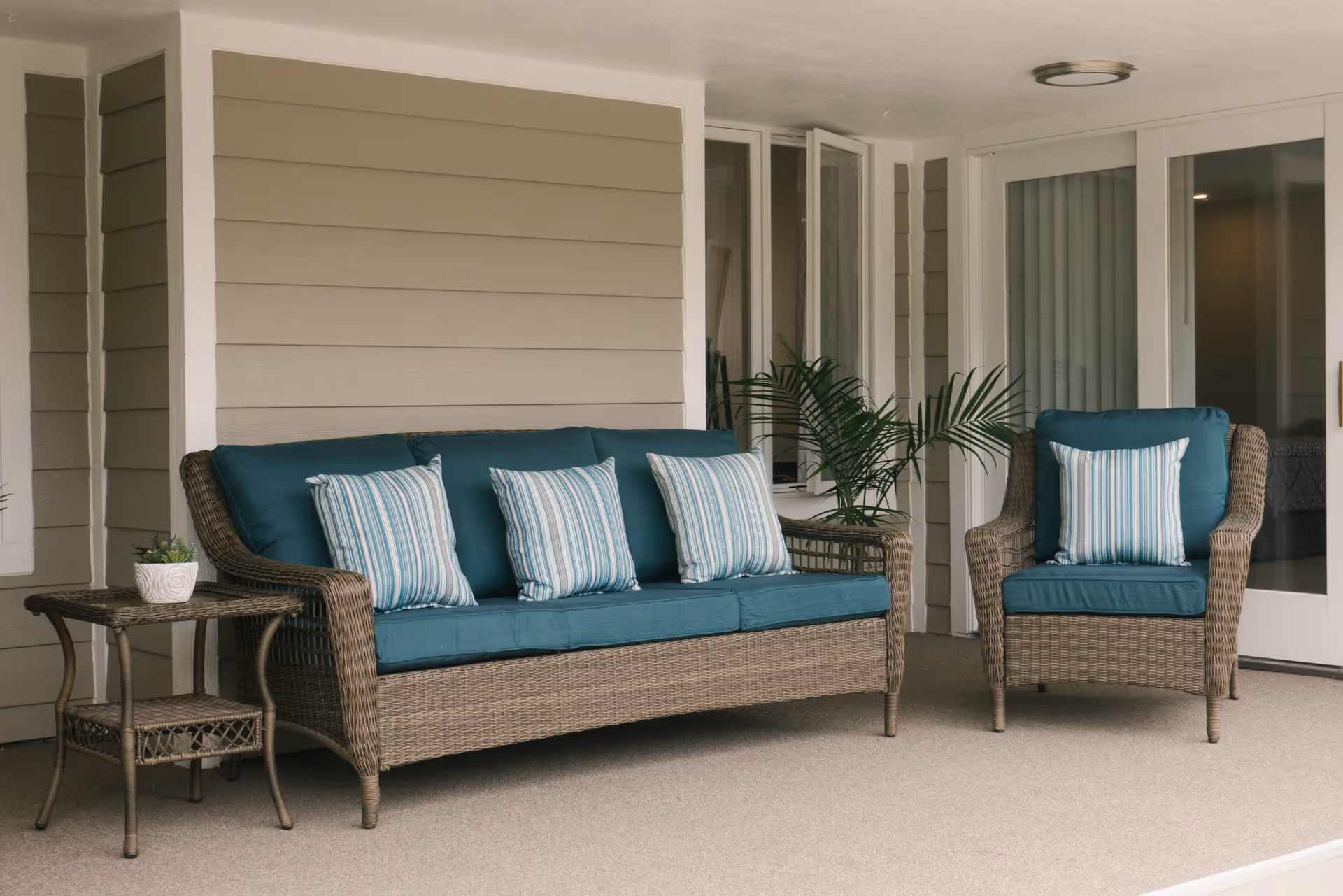 A covered outdoor patio area with a wicker sofa and armchair, both with teal cushions and striped throw pillows. There is a small wicker side table with a potted plant on it, and a green leafy plant in the corner near glass doors and windows.