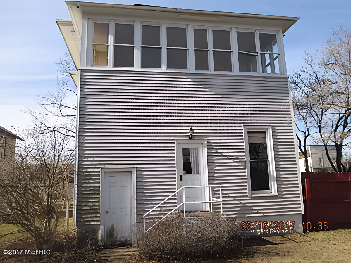 Front exterior of a two-story light-gray siding house with an enclosed upper porch, a central door with steps and railing, and bare trees nearby.