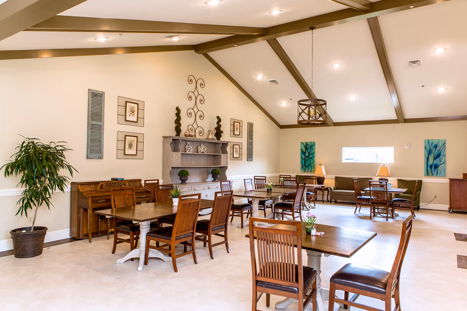 Spacious dining/common room with multiple wooden tables and chairs, a piano, decorative shelving, and a high beamed ceiling.