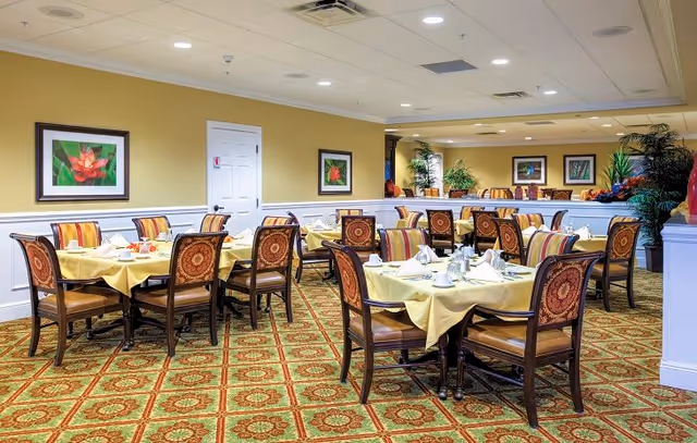 A dining room with multiple tables covered in yellow tablecloths, each set with white napkins, cups, and plates. The chairs have patterned upholstery with a floral design. The room has a patterned carpet, yellow walls with white wainscoting, framed floral artwork, and several potted plants.