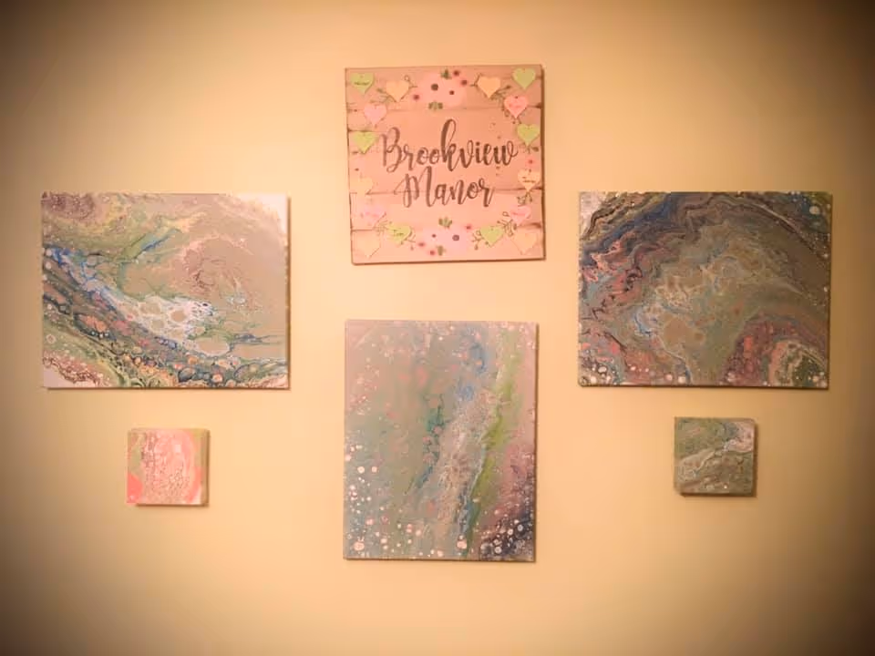 A beige wall with five pieces of artwork arranged in a pattern. The top center piece is a wooden plaque with the text 'Brookview Manor' surrounded by small heart-shaped notes. The other four pieces are abstract paintings with swirling colors including green, pink, blue, and white.
