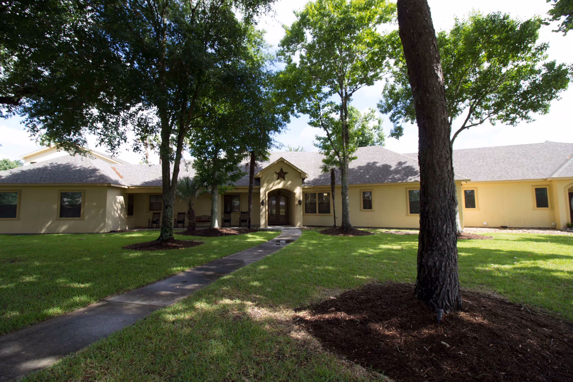 Front exterior view of a single-story senior living facility building with a beige facade, a central entrance with double doors, a star decoration above the entrance, surrounded by green grass and several tall trees providing shade.