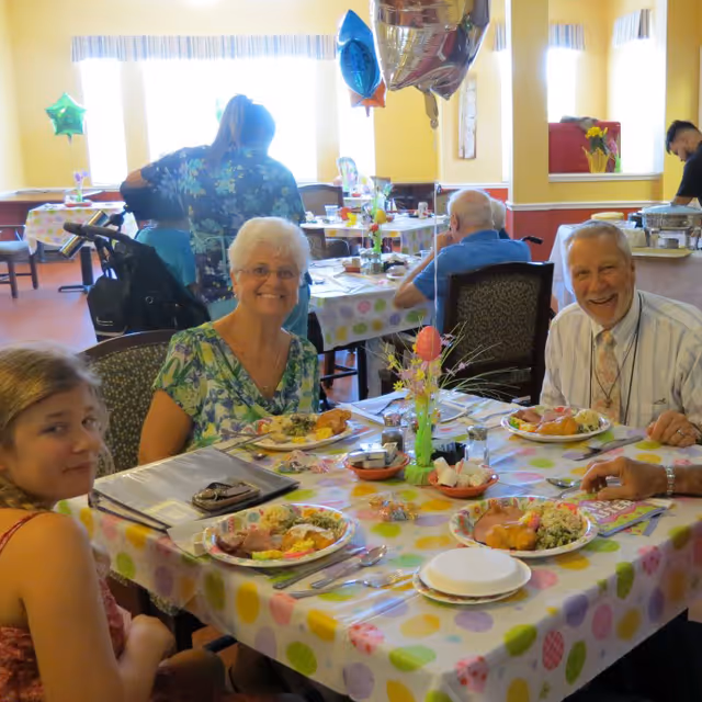 Several smiling older adults and a child seated around a decorated table with plates of food in a brightly lit dining room.