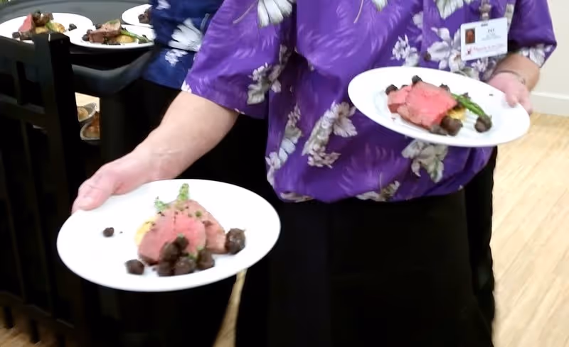 A person wearing a purple floral shirt and a name badge is holding two plates of food, each with slices of roast beef, asparagus, and mushrooms. The background shows more plates of food on a dark surface.