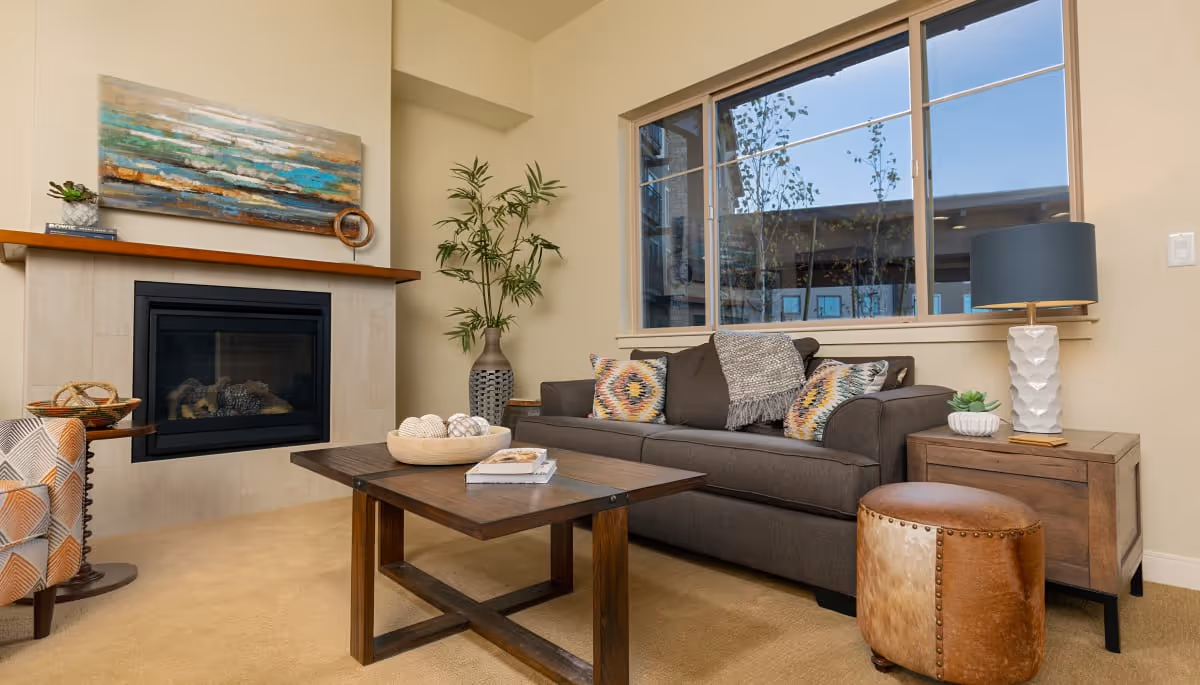 A cozy living room with a dark gray sofa adorned with colorful patterned pillows and a gray throw blanket. In front of the sofa is a wooden coffee table with decorative items and books. To the right of the sofa is a wooden side table with a white textured lamp and a small plant. A brown leather ottoman is placed nearby. On the left side, there is a fireplace with a wooden mantel, decorated with a small plant and a circular ornament. Above the fireplace hangs a large abstract painting with blue, green, and beige tones. A tall potted plant is positioned in the corner near a large window showing an outdoor view.