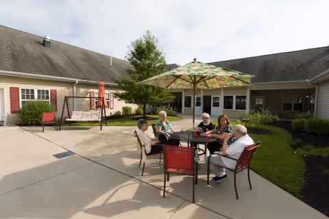 Five elderly people sitting around a round outdoor table with a large umbrella, enjoying drinks and conversation in a courtyard area of a senior living facility. The courtyard has concrete pavement, green grass, shrubs, and a swing bench. The building surrounding the courtyard has beige siding and red window shutters.