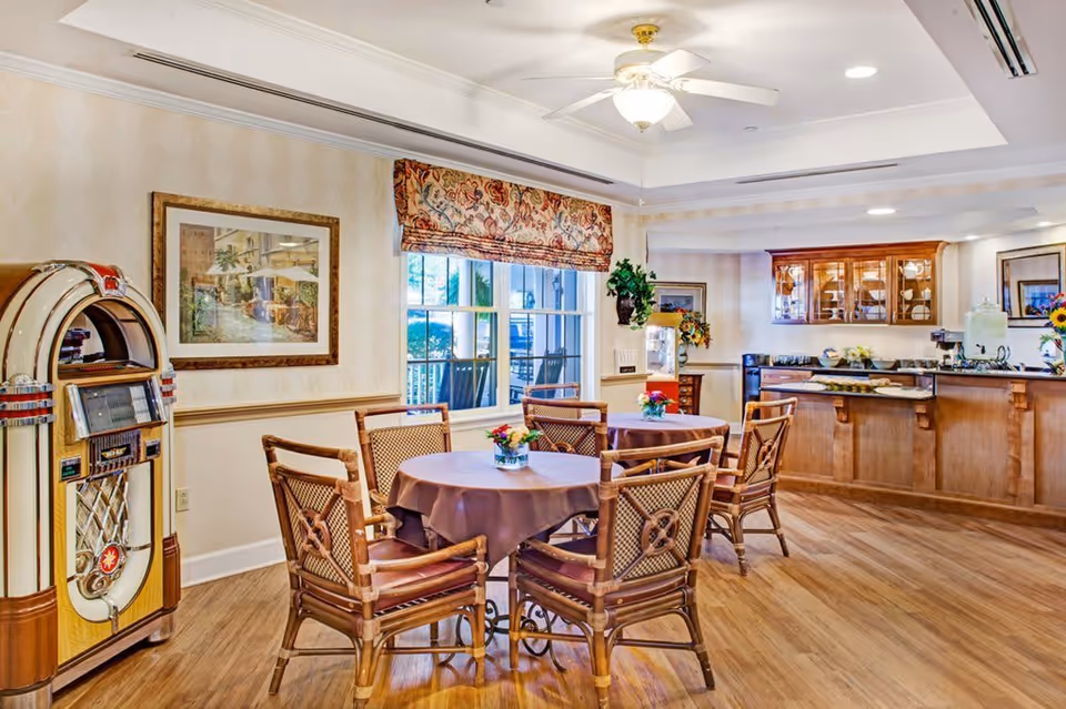 A cozy dining area in an assisted living facility featuring round tables with brown tablecloths and wooden chairs. A vintage jukebox stands against the wall on the left. The room has wooden flooring, a large window with a floral valance, framed artwork on the walls, and a counter area with cabinets and refreshments in the background.