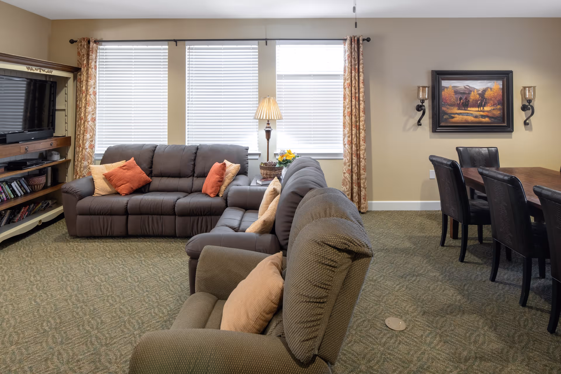 A cozy living room area with two brown sofas and a green recliner chair, each with beige and orange throw pillows. There is a wooden entertainment center with a TV and books on the left side, three large windows with blinds and floral curtains in the background, and a floor lamp between the sofas. On the right side, there is a dining table with six black chairs, a framed painting on the wall, and two wall-mounted candle holders.
