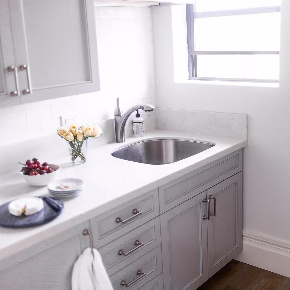 Bright modern kitchen countertop with a stainless-steel sink, gray cabinetry, a vase of pale roses and small bowls beside a window.
