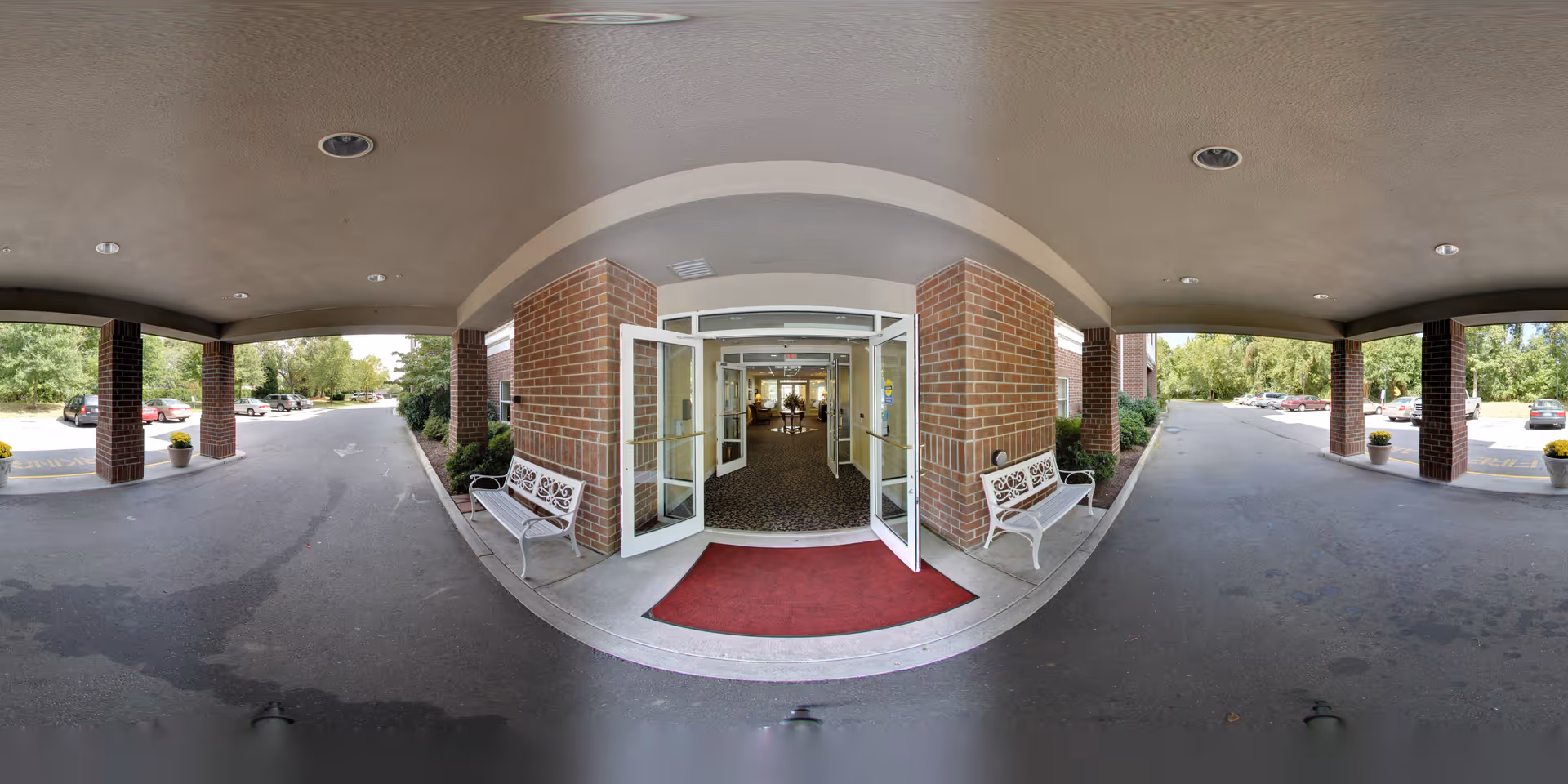 Covered entrance area to a building with double glass doors open, two white benches on either side, a red doormat, and a parking lot with cars and greenery visible in the background.