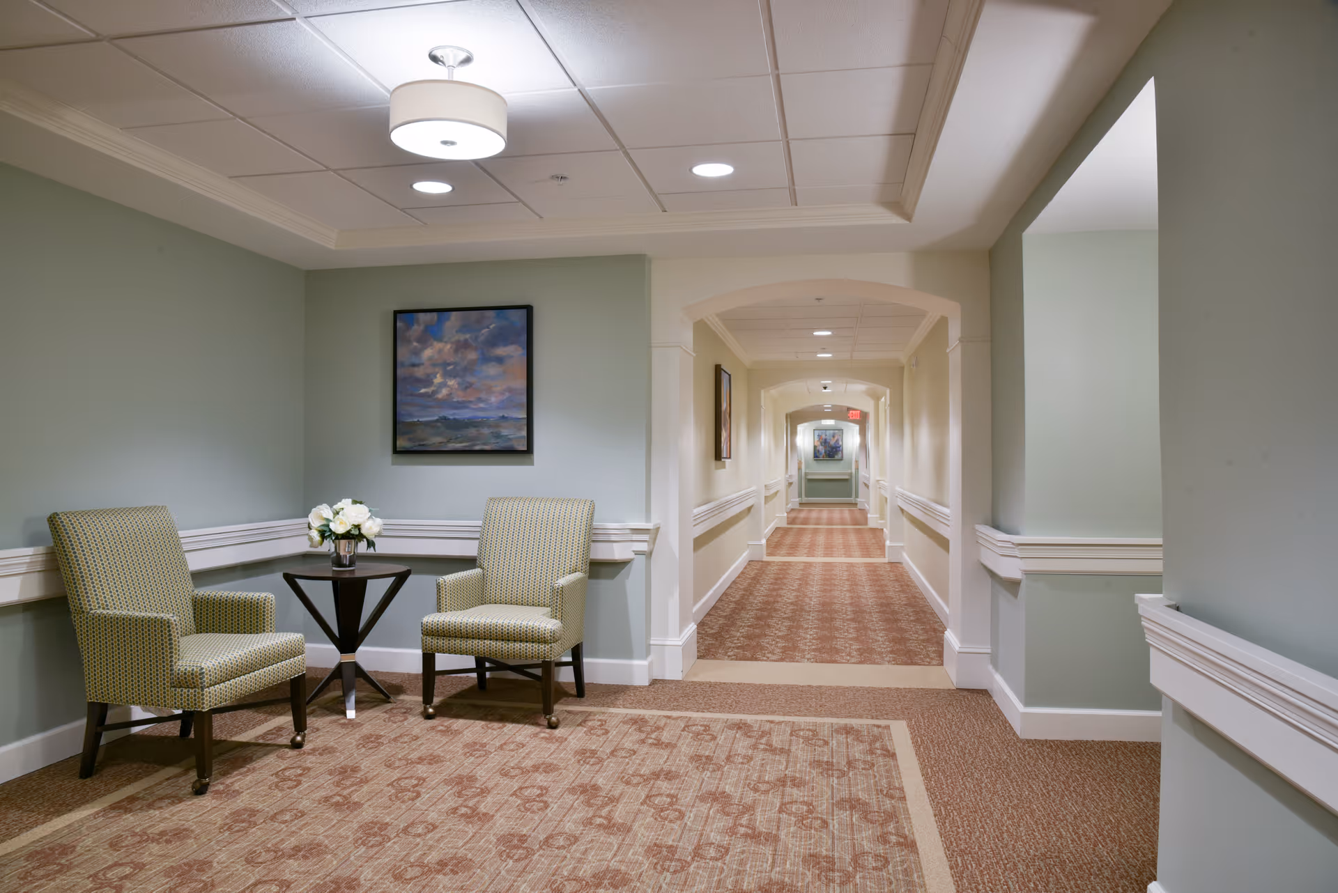 A well-lit hallway in a senior living facility with light green walls, patterned carpet, and white trim. Two upholstered chairs and a small round table with a vase of white flowers are placed in a corner. A framed painting hangs on the wall above the chairs. The hallway extends into the distance with additional artwork visible on the walls.