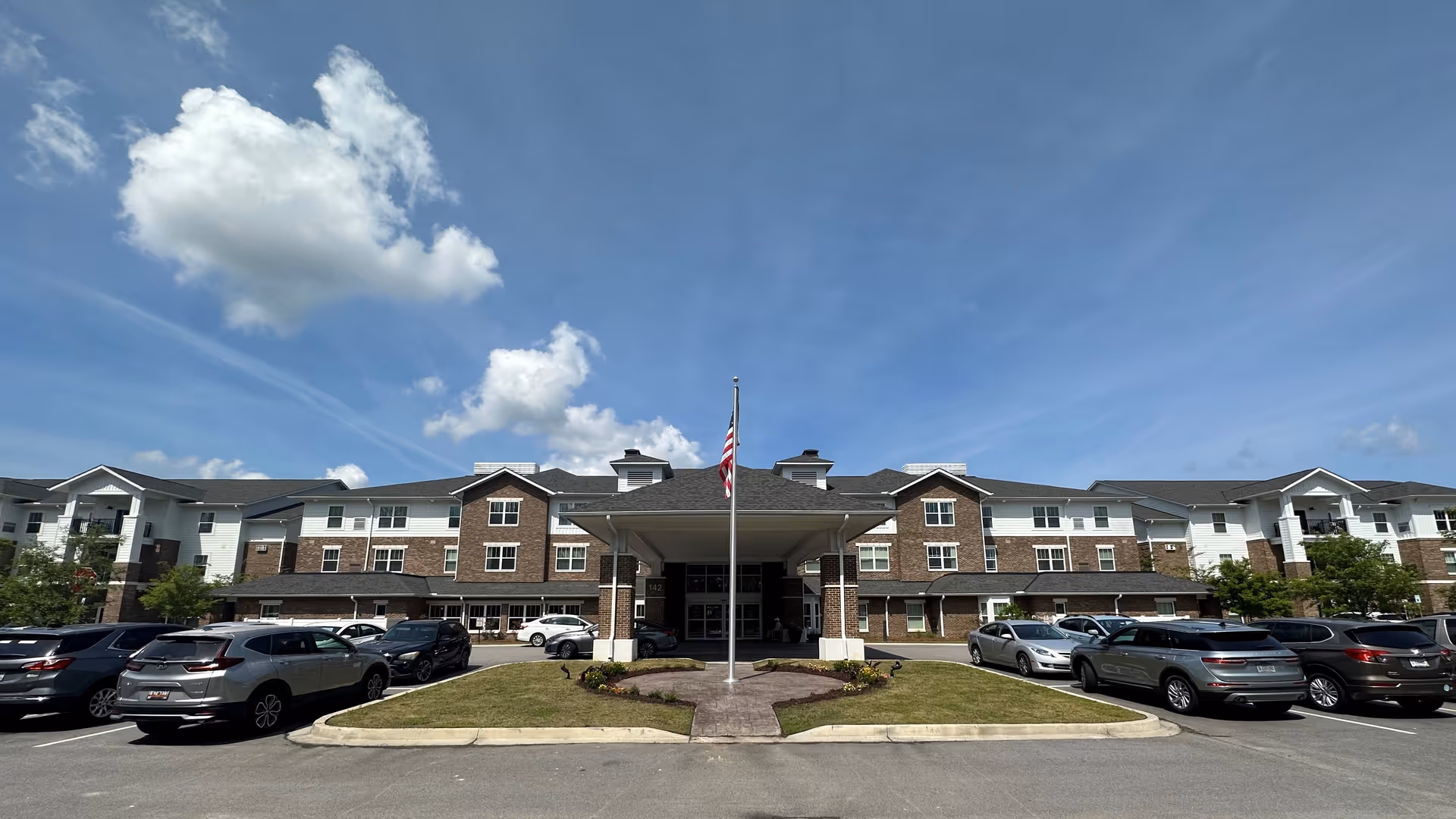 Front entrance of a multi-story senior living building with a covered porte-cochere, flagpole, and parked cars under a blue sky.