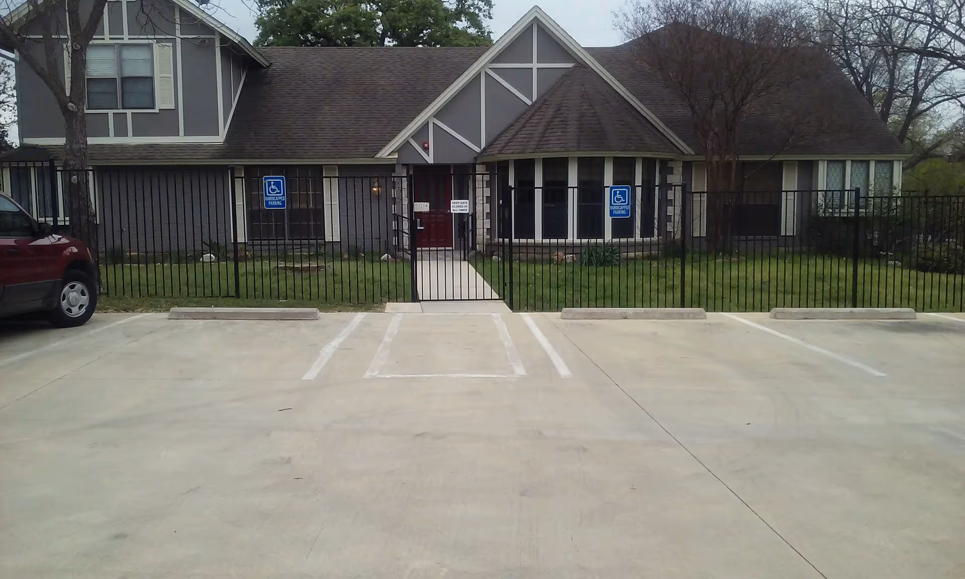 Exterior view of a single-story assisted living facility with a gray facade, white trim, and a dark shingled roof. There is a black metal fence with a gate in front of the entrance, which has a red door. Two handicapped parking signs are visible on the fence, and there is a red vehicle parked on the left side of the image. The area in front of the building is a concrete parking lot with marked parking spaces.