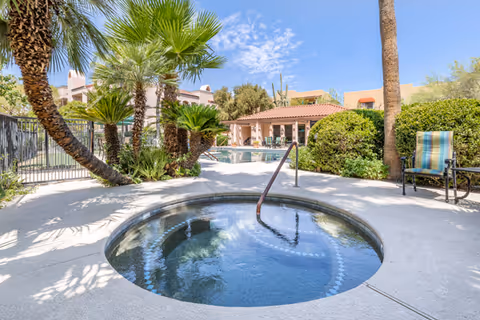 Outdoor area at Brookdale Santa Catalina featuring a circular hot tub in the foreground, surrounded by palm trees and other greenery. In the background, there is a swimming pool, a building with a tiled roof, and a chair with a striped cushion under a clear blue sky.